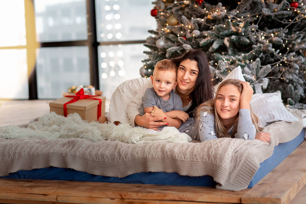 Gelukkige vrolijke familie in de buurt van kerstboom. Moeder en kinderen die 's morgens een pyjama dragen en zich vermaken in de buurt van een boom. Vrolijk kerstfeest en fijne feestdagen concept - Foto, afbeelding