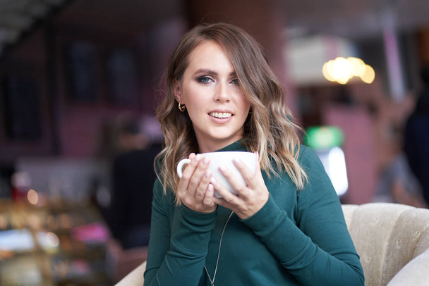 Een mooie vrouw met een dameskapsel en een kop koffie in haar hand zit in een café. Hij kijkt met een glimlach naar de camera. - Foto, afbeelding