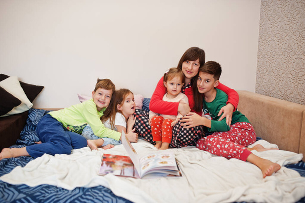 Happy big family is having fun together in bedroom. Large family morning concept. Mother with four kids wear pajamas read book in bed at home. - Фото, изображение