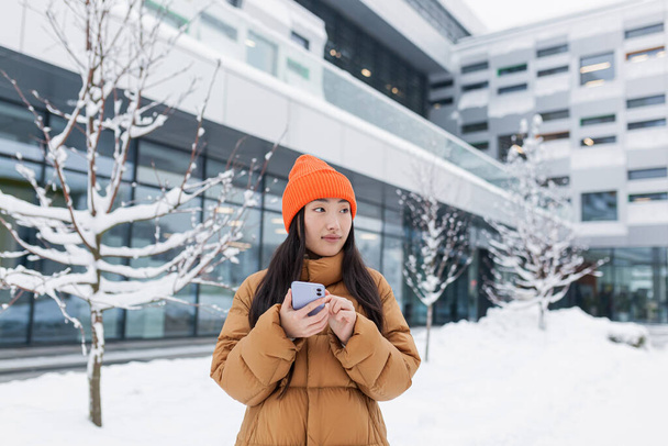 Beautiful Asian girl uses the phone, online dating, makes a choice, walks on a winter snowy day - Valokuva, kuva