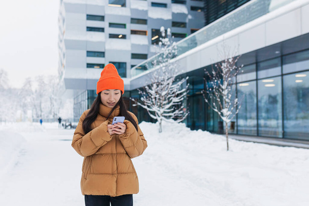 Jeune belle étudiante asiatique, femme millénaire utilise le téléphone, marche par une journée enneigée, hiver dans une veste chaude et chapeau près du campus universitaire - Photo, image