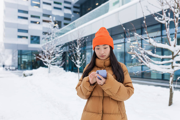 Nuori kaunis aasialainen opiskelija, tuhatvuotinen nainen käyttää puhelinta, kävelee lumisena päivänä, talvella lämpimässä takissa ja hatussa yliopiston kampuksen lähellä. - Valokuva, kuva