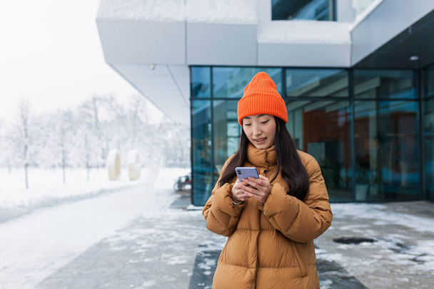 Young beautiful Asian student, millennial woman uses the phone, walks on a snowy day, winter in a warm jacket and hat near the university campus - Foto, afbeelding