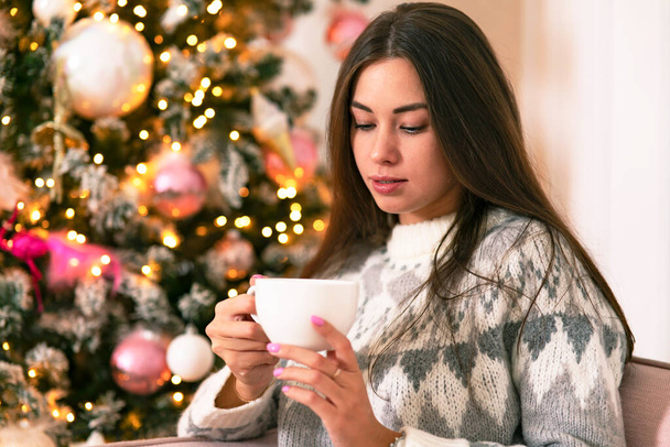 Beautiful girl with a mug in her hands greets the new year and christmas by the tree. Girl at home, watching a drink, cozy - Zdjęcie, obraz