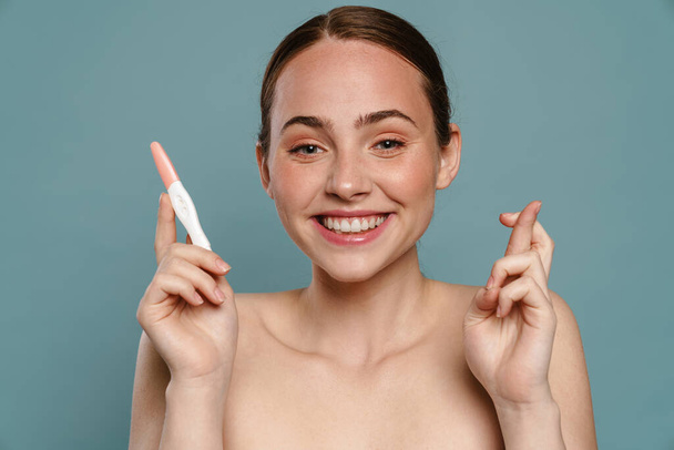 Half-naked ginger woman gesturing and showing pregnancy test isolated over blue background - Foto, afbeelding