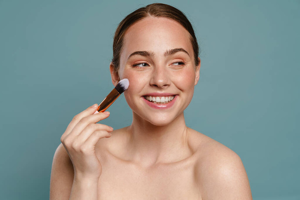 Half-naked ginger woman smiling and using cosmetic brush isolated over blue background - Foto, afbeelding
