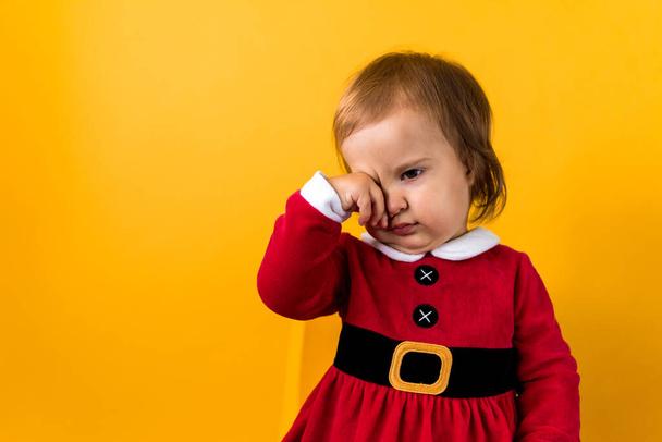 Banner Portraite Cute Happy Cheerful Chubby Baby Girl in Santa Suit Sleepy Rubs Eyes At Yellow Background. Child Play Christmas Scene Celebrating Birthday. Kid Have Fun Spend New Year Time Copy Space - Foto, Imagen