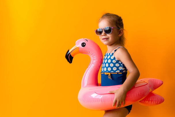 happy little child swimmer sport girl wear sun glasses swimsuit, Kid Inflatable swimming ring Pink Flamingo point to side away, isolated orange background. childhood, hot summer vacation hobby concept - Fotografie, Obrázek