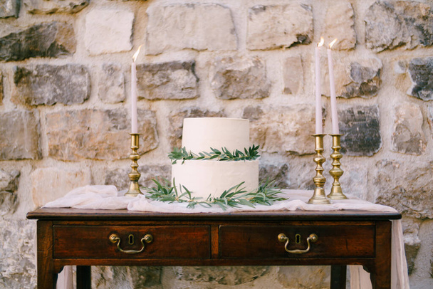 Gâteau de mariage superposé décoré de branches vertes se dresse sur une table en bois près d'un mur de pierre - Photo, image
