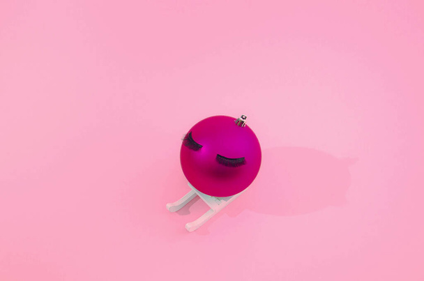 A fuchsia Christmas bauble with lashes on a white sleigh on pink background. Funny trending minimal creative concept for New Year or Christmas card or advertisement or banner. - Fotó, kép