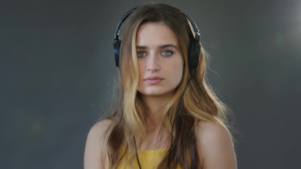 Stúdió portré szürke háttér fiatal gyönyörű komoly lány diák visel fejhallgató fej töprengő modell Y-generációs szőke nő fülhallgató zenét hallgat audio dal nézi kamera - Felvétel, videó