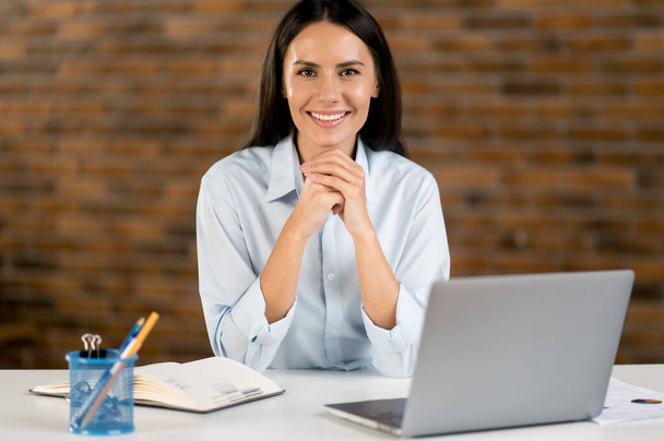 Portrait young adult successful beautiful caucasian business woman, office worker, manager, business consultant, dressed in formal shirt, sitting at her workplace with laptop, looking at camera, smile - Photo, Image