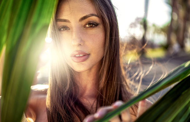Zbliżenie portret piękna zmysłowej brunetki z długimi włosami i brązowymi oczami. Dziewczyna pozowanie na zewnątrz w słoneczny dzień, patrząc na aparat przez zielone liście - Zdjęcie, obraz