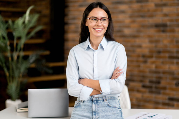 Portrait successful cute confident beautiful young adult caucasian businesswoman or broker standing near work desk in office dressed in stylish clothes, arms crossed, looking to side, smiling - Photo, image