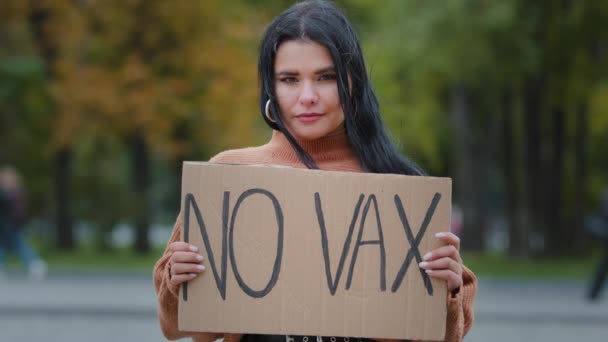 Young confident hispanic woman holding cardboard sign no vax protesting against vaccination outdoors serious girl standing on street demonstrates protest immunization covid19 stop untried vaccine - Footage, Video