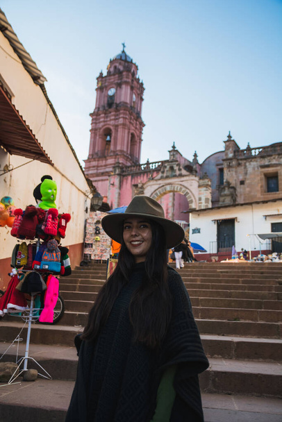 Adventurous girl, exploring the Magical Town of Tlalpujahua, Mexico. She smiles as she admires its streets and churches of classic Mexican architecture wearing a black poncho and a green hat. - Valokuva, kuva