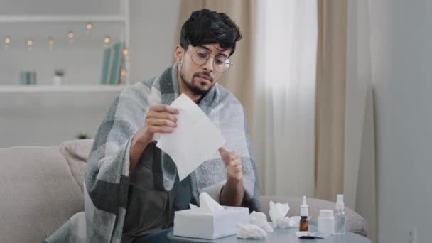 Nuori sairas kaveri arabialainen silmälasipäinen mies istuu kotona sohva peitetty huopa ruudullinen kärsii kylmästä sairaudesta kausiluonteinen allergia virus puhaltaa vuotava nenä paperiin lautasliina tunne huonovointisuus terveysongelmia - Materiaali, video