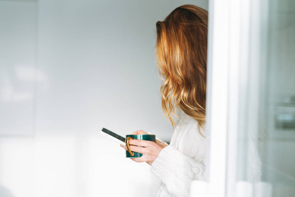 Mladý dospělý čtyřicet let krásná blondýnka s dlouhými vlasy pomocí smartphone a pití ranní čaj v kuchyni doma - Fotografie, Obrázek