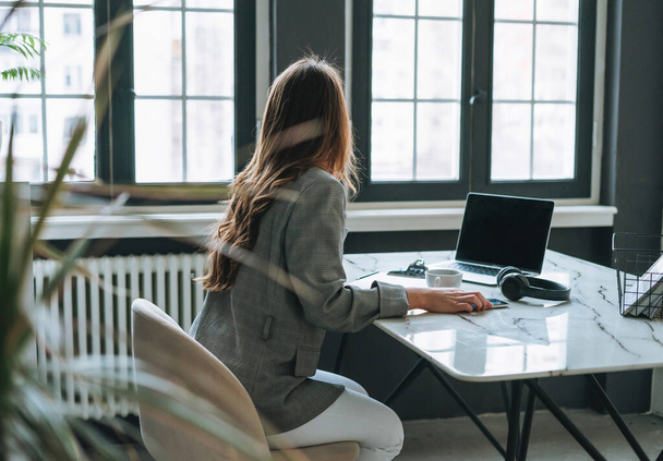Gri takım elbiseli uzun saçlı esmer kadın modern ofiste dizüstü bilgisayarla çalışıyor. - Fotoğraf, Görsel