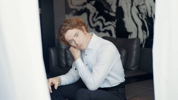 A man with a white dress shirt. Action. A man with red hair is sitting on the sofa and adjusting the collar of his clothes against a background of light black wallpaper and a bright white curtain in - Zdjęcie, obraz