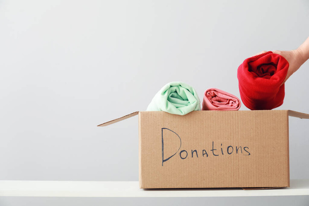 Vrouw zet donatie kleding in doos op plank tegen lichte achtergrond - Foto, afbeelding