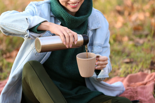 Young woman sitting on plaid and pouring tea from thermos in autumn park - Photo, Image