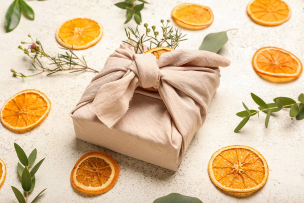 Composizione con confezione regalo avvolta in tessuto, rami vegetali e fette di arancia secca su fondo chiaro - Foto, immagini