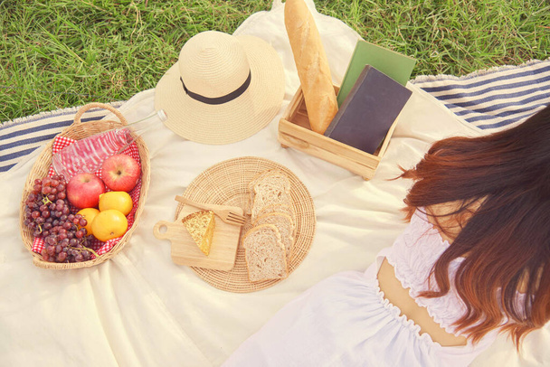 Piknik konsepti. Uzun kahverengi saçlı, rahat beyaz elbiseli kızın arkası. Piknik örtüsüne uzanıp kitap okumak.. - Fotoğraf, Görsel