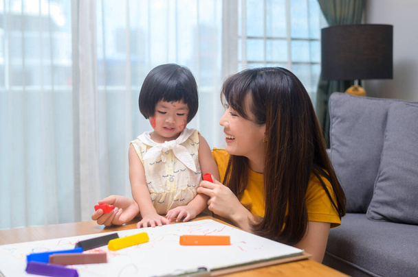 Μια νεαρή μαμά βοηθά την κόρη ζωγραφίζει με χρωματιστά μολύβια στο σαλόνι στο σπίτι. - Φωτογραφία, εικόνα