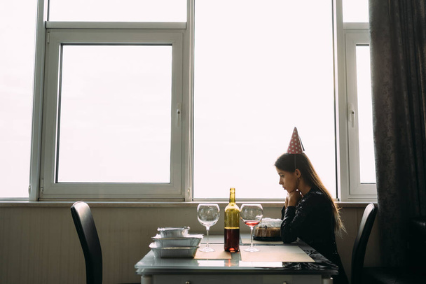 triste fille d'anniversaire solitaire, dame assise avec gâteau à la maison près de la fenêtre, fille d'anniversaire malheureuse près de la fenêtre. Fête solitaire, concept d'anniversaire à distance - Photo, image
