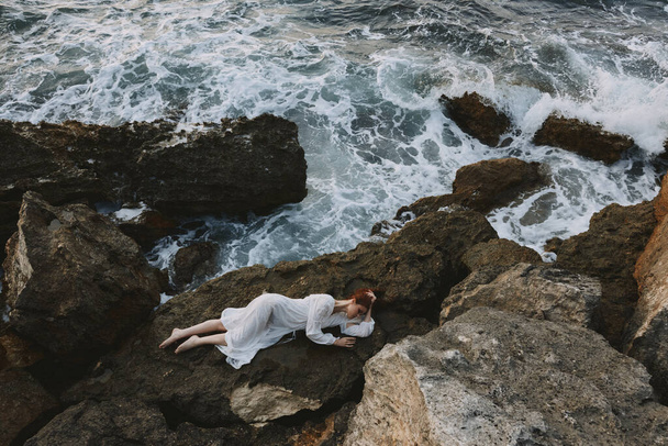 beautiful woman in long white dress wet hair lying on a rocky cliff nature landscape - Photo, Image