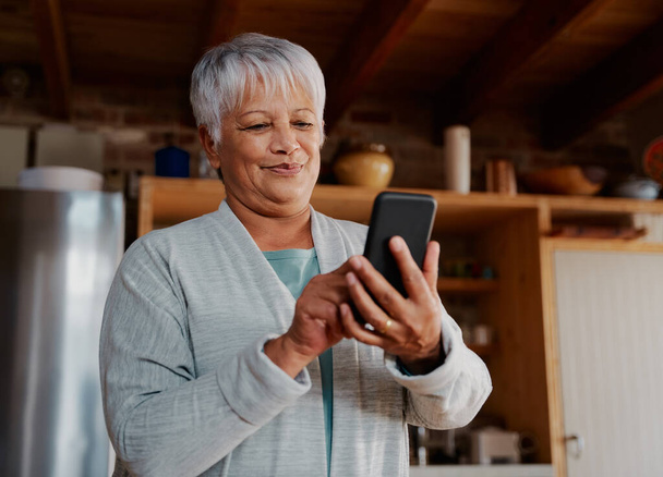 Content multi-cultural elderly female typing a message on smartphone while standing in modern kitchen holding morning coffee. - Photo, image