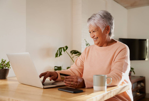 Elderly multi-ethnic female happily making online payment on laptop, sitting at kitchen counter online banking. - Фото, изображение