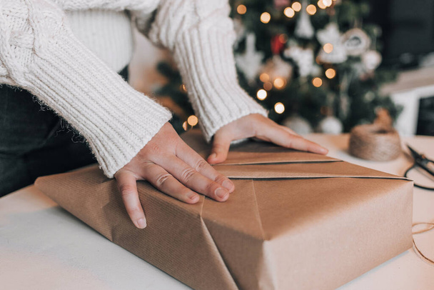 Midsection of woman wearing cozy thick sweater, wrapping christmas present in kraft paper. Christmas tree with bokeh lights in background. - Foto, afbeelding