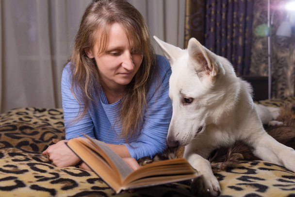 A woman lying on bed and reading a book with her companion white swiss shepherd dog. Love, togetherness and pets indoors. - Fotó, kép