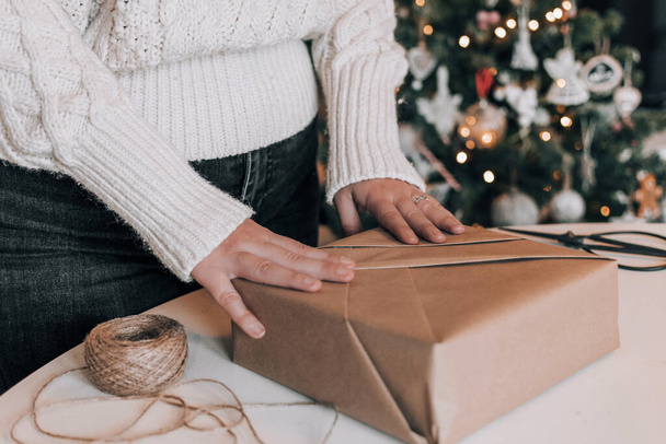 Midsection of woman wearing cozy thick sweater, wrapping christmas present in kraft paper. Christmas tree with bokeh lights in background. - Foto, Bild
