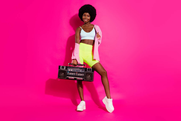 Photo of sweet pretty dark skin woman dressed crop top smiling listening boombox isolated pink color background - Foto, afbeelding