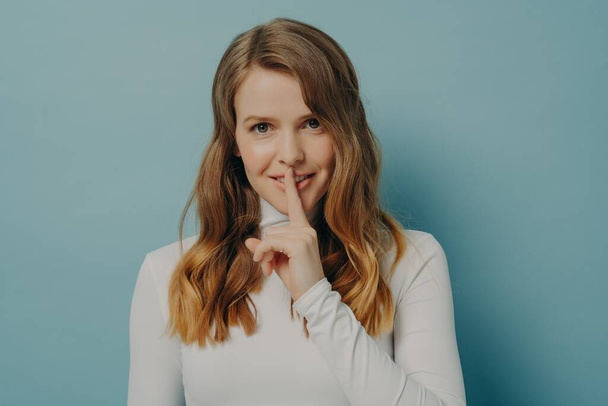 Smiling young millennial woman holding index finger on her lips over blue studio wall background - Photo, Image