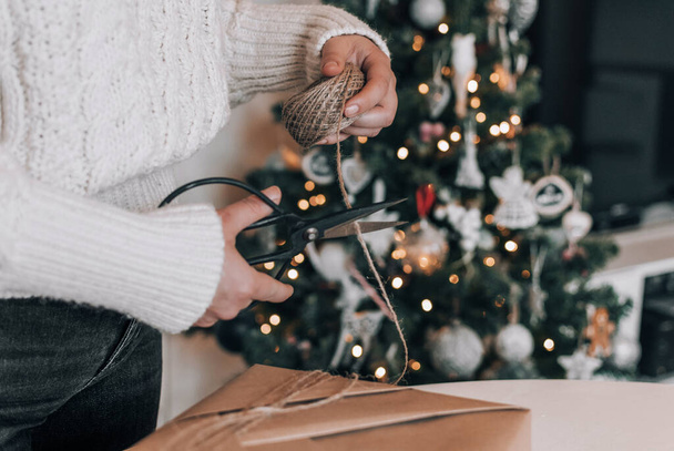 Midsection of woman wearing cozy thick sweater, wrapping christmas present in kraft paper. Christmas tree with bokeh lights in background. - Fotoğraf, Görsel