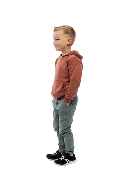 Smiling boy stands holding his hands in his pockets. Positiveness and happiness. The guy in jeans and a brown sweater. Isolated on white background. Vertical. - Photo, Image