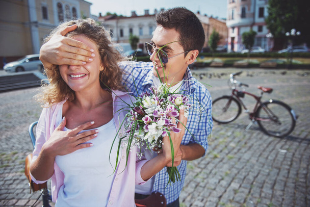 Guess who? Young man closed eyes his girlfriend making surprise with flowers while both standing at the city street. - Photo, Image