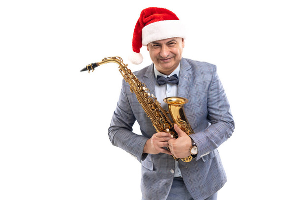 Funny musician wears in Santas hat holds saxophone while showing thumb up on studio background - Foto, Bild