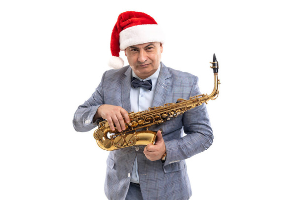 Sceptical musician man wears in Santas hat holds saxophone over studio background - Photo, image