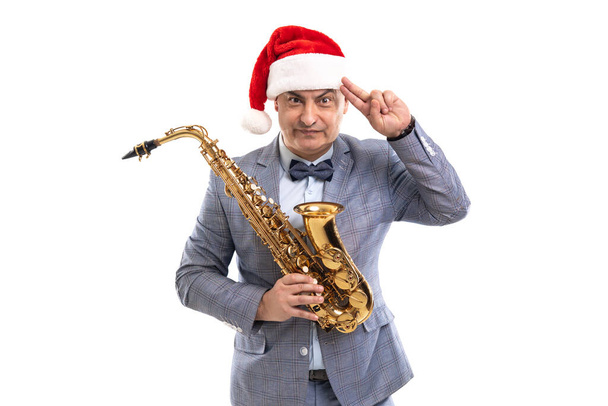 Sceptical man wears in Santas hat holds saxophone while raising hand to head on studio background - Zdjęcie, obraz