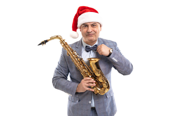 Sceptical man wears in Santas hat holds saxophone while straightening bow tie on studio background - Photo, image