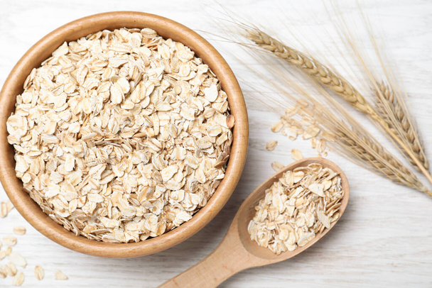 Oatmeal, bowl, spoon and spikelets on white wooden table, flat lay - Foto, imagen
