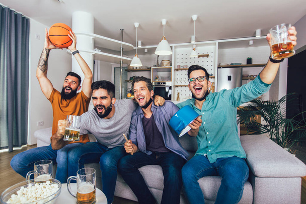 Feliz macho amigos vistiendo cara negra máscara protectora viendo baloncesto en casa - Foto, imagen