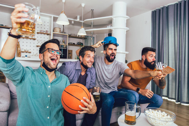 Happy male friends  wearing black face protective mask watching basketball at home - Fotó, kép