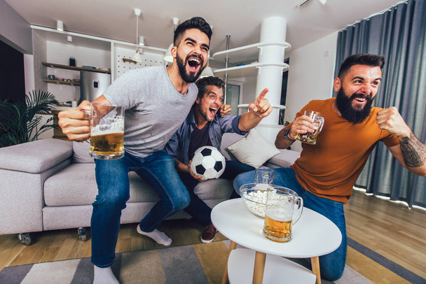 Πολύ ενθουσιασμένοι φίλοι διασκεδάζουν βλέποντας ποδοσφαιρικό αγώνα στο σπίτι, σε εσωτερικούς χώρους. Φιλία, leasure, αθλητισμός, ανάπαυση, έννοια σπίτι κόμμα - Φωτογραφία, εικόνα