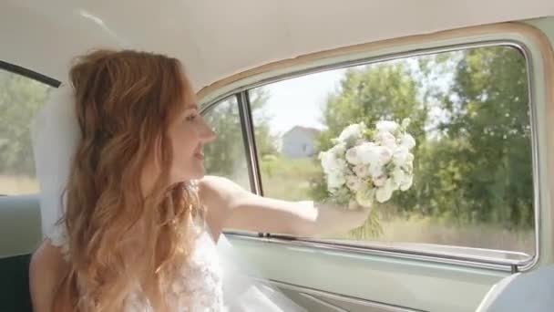 Eine charmante junge Braut im Schleier streckte ihre Hand mit einem Brautstrauß im Fenster eines in der Natur fahrenden Retro-Autos aus. Zeitlupe - Filmmaterial, Video